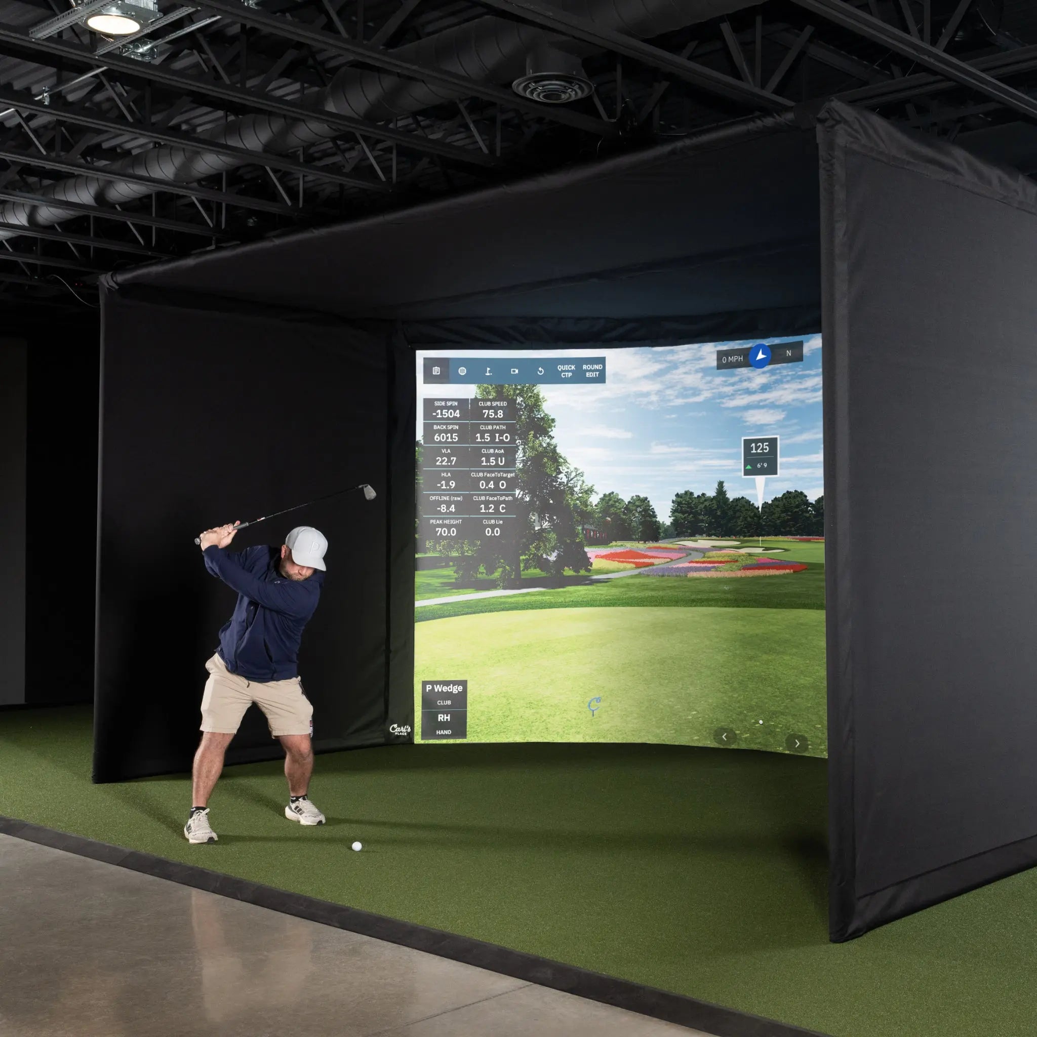 A golfer in a blue shirt and white cap is preparing to swing a club in front of a large curved golf simulator screen displaying a realistic course landscape and performance metrics.