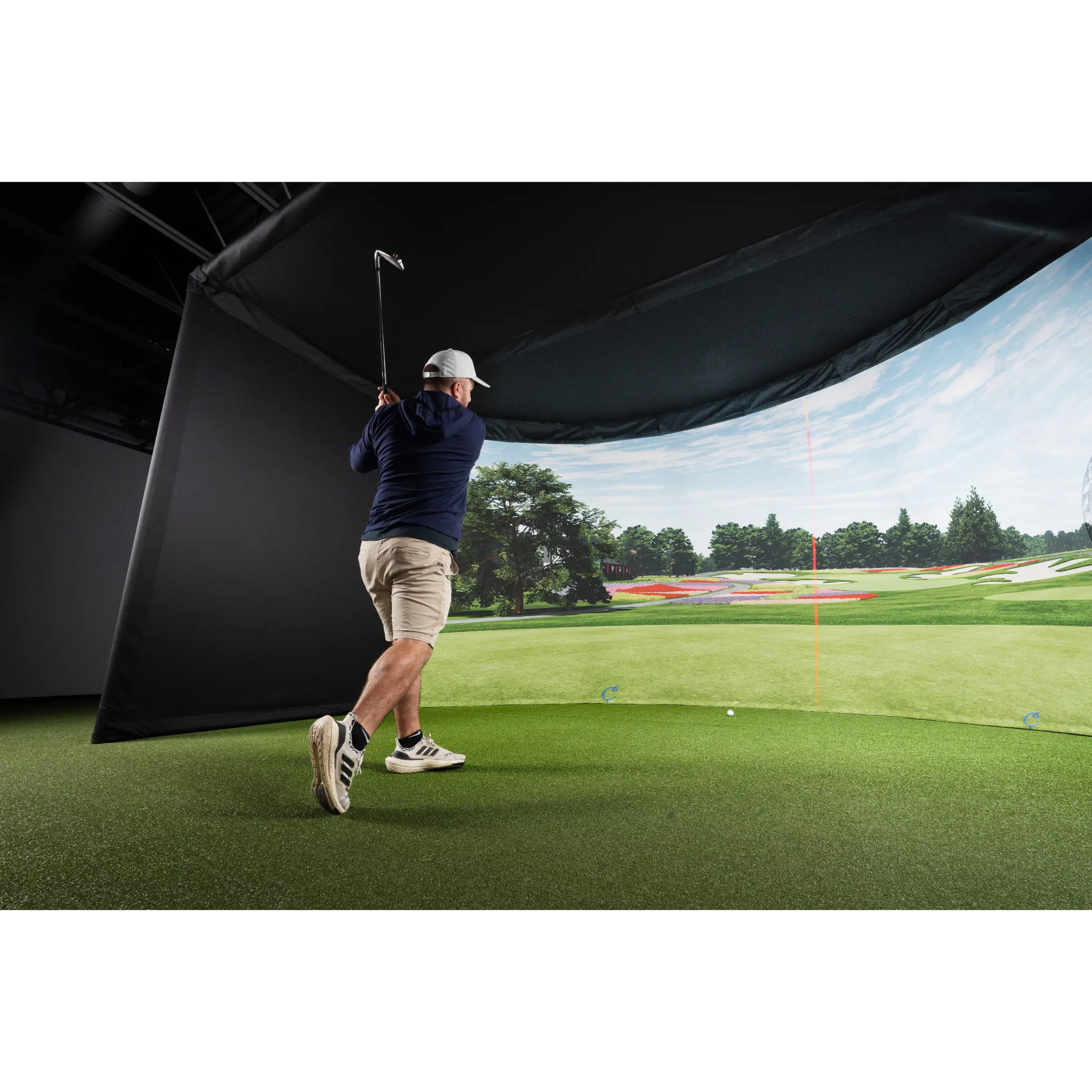 A golfer swings a club in an indoor setting featuring a curved simulator enclosure. The backdrop displays a realistic golf course scene with lush green grass and a blue sky, enhancing the immersive golfing experience.