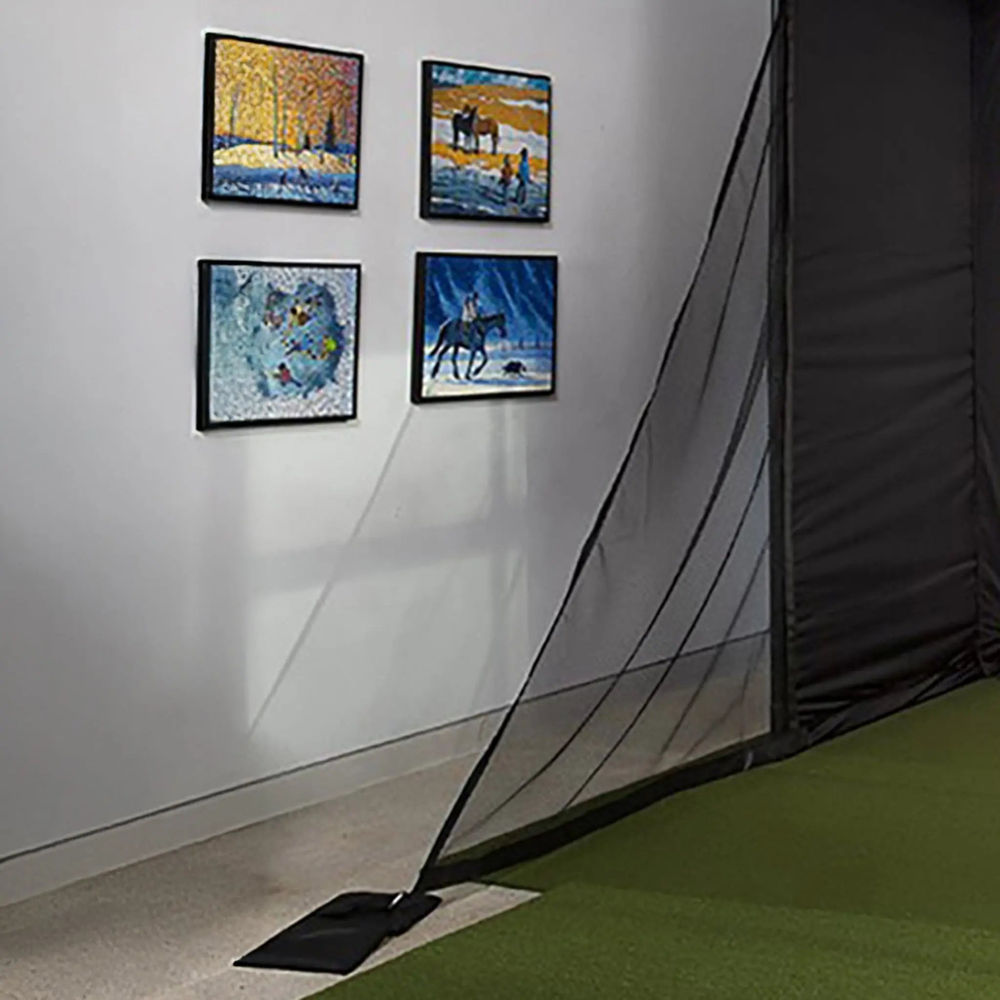 A golf simulator setup featuring a black enclosure with a semi-transparent impact screen that reaches the floor. Four framed artworks are hanging on the wall, adding a decorative touch to the space.