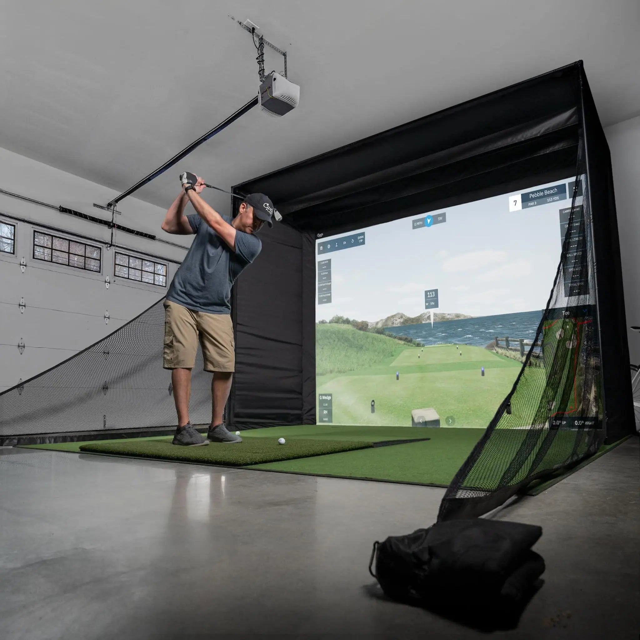 A man stands in a garage, taking a golf swing towards a large impact screen displaying a scenic golf course. The setup features a green turf mat and a black enclosure, creating an immersive indoor golfing experience.