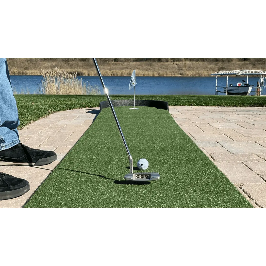Person putting a golf ball on the Big Moss Commander Patio Series 215 green near a lakeside patio