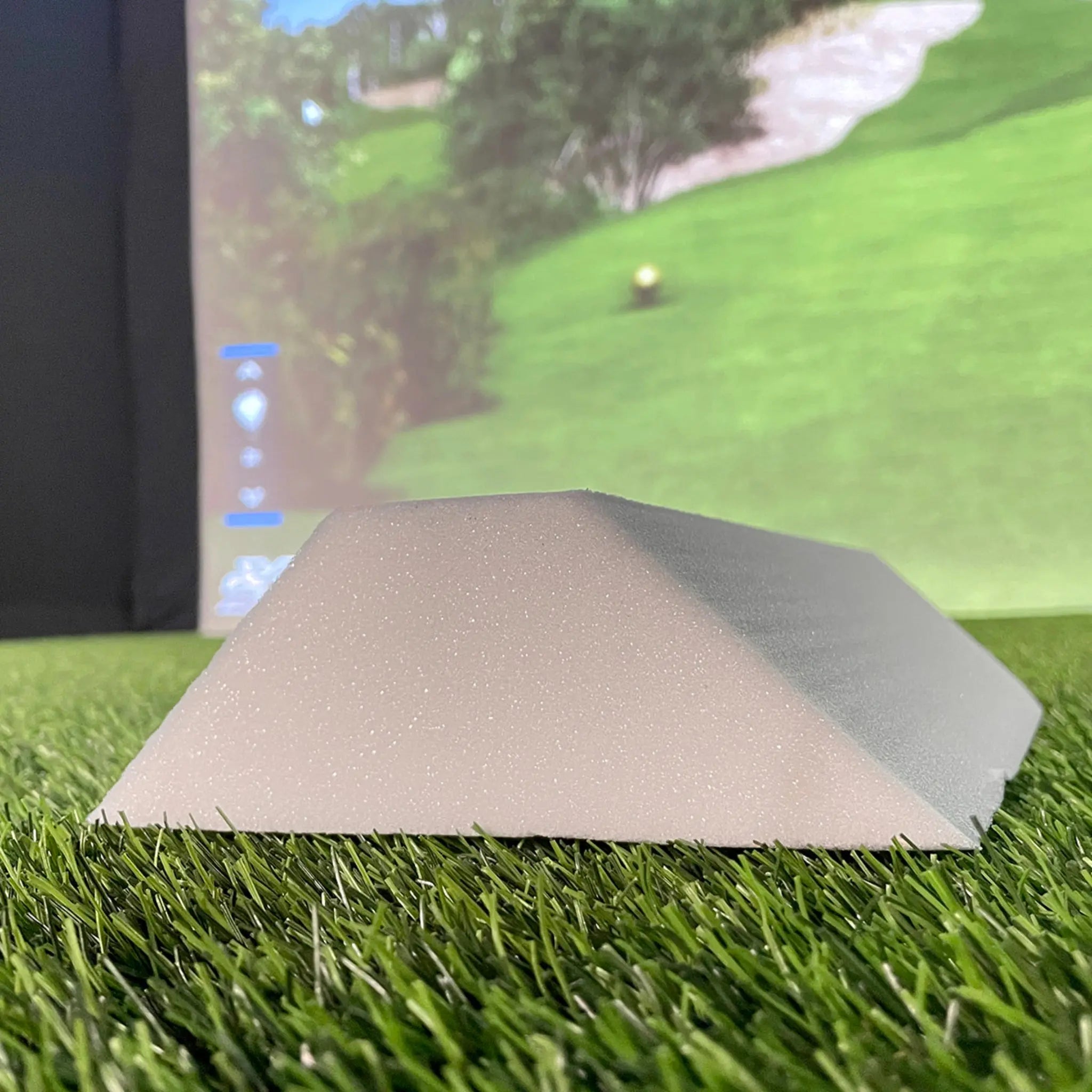 A close-up view of a triangular foam piece resting on artificial grass, with a blurred golf simulation background featuring a green landscape and a golf ball in the distance. The foam appears to be part of a golf simulator setup.