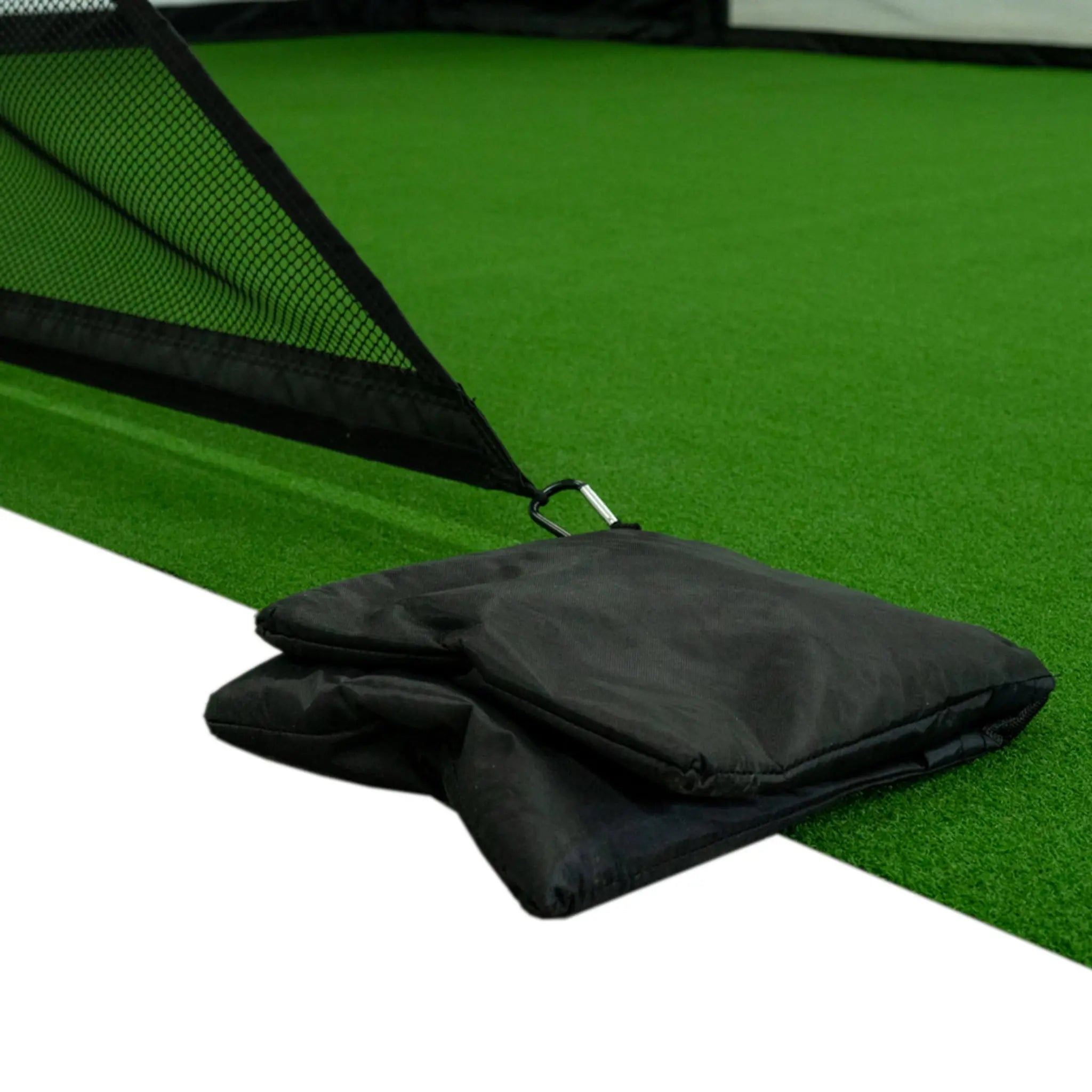 A close-up view of a black nylon weight bag designed to secure a golf simulator enclosure on a vibrant green artificial turf surface. The bag is folded and positioned near the edge of the enclosure, highlighting its durable construction.