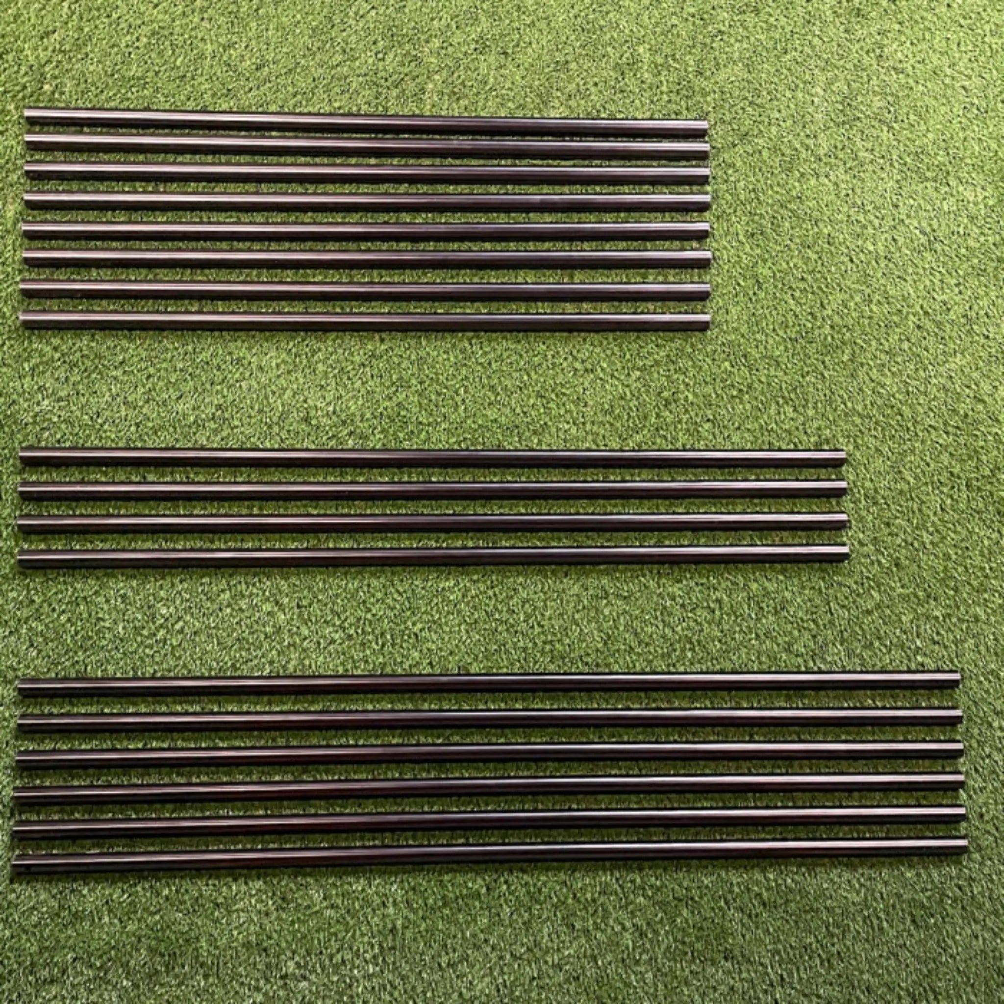 A close-up view of several black metal rods arranged in parallel lines on a green synthetic grass surface. The rods vary in length and are neatly organized, showcasing their sleek design intended for construction or assembly purposes.
